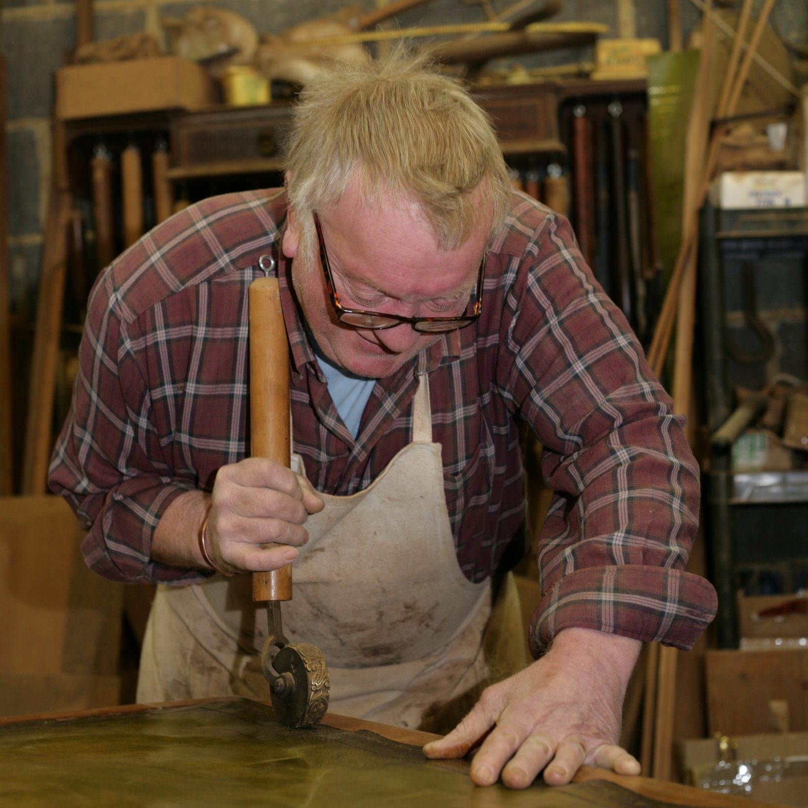 An antique restorer uses a hand-roller to incise a decorative motif around the edge of an antique desk's inset leather writing surface.  Gold leaf is then often used to highlight the design.  